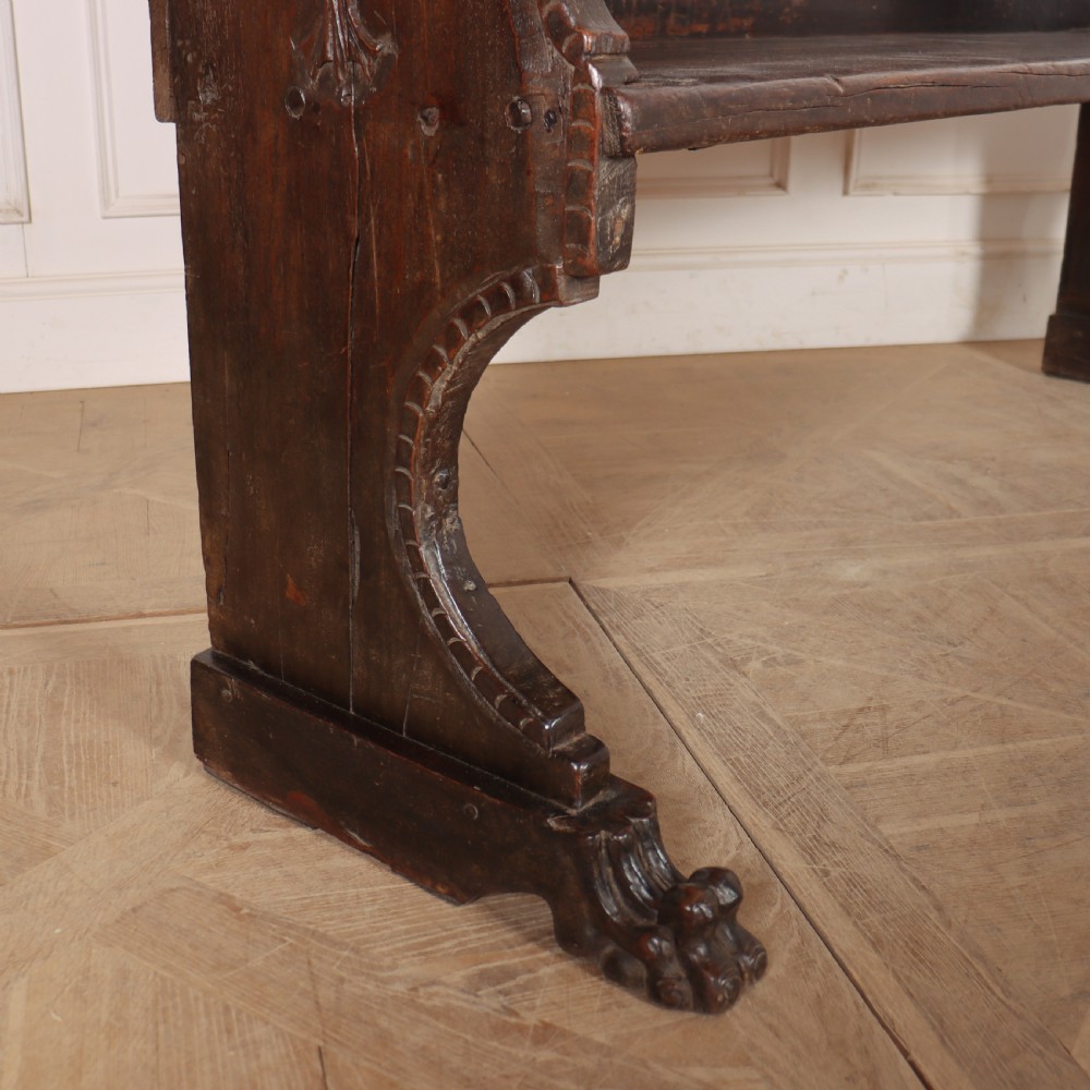 A dark brown carved walnut bench with a panelled backrest featuring carved rosette medallions and detailed carved feet. View X