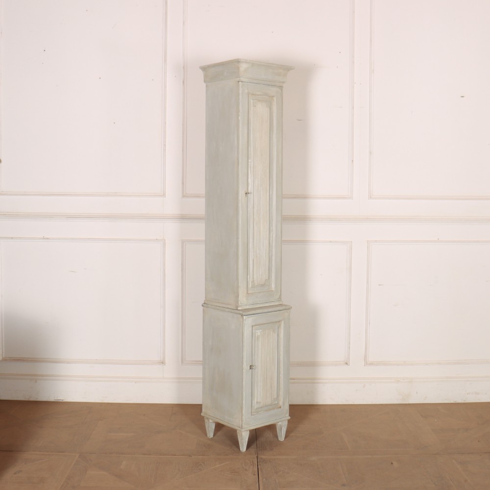 A tall, narrow Gustavian style two-tier painted wooden cabinet in pale grey with fluted panel doors and tapered feet. View II