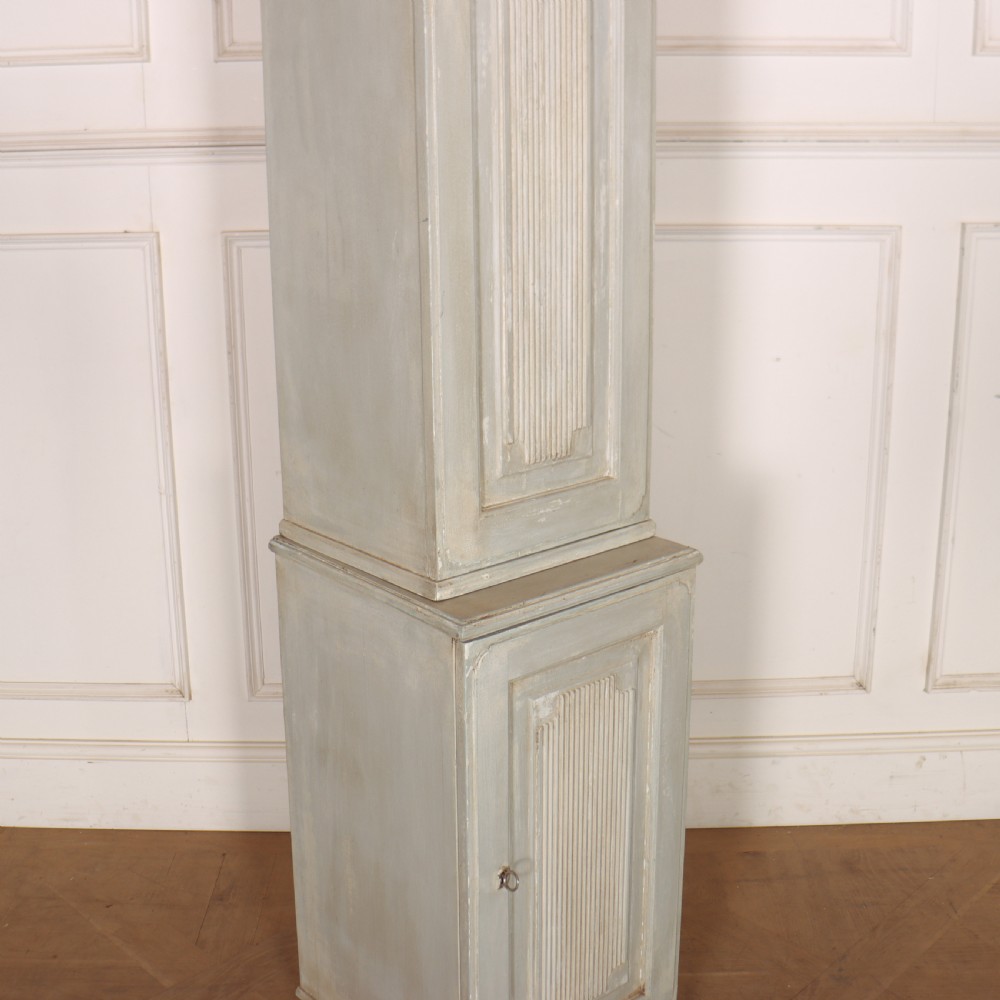A tall, narrow Gustavian style two-tier painted wooden cabinet in pale grey with fluted panel doors and tapered feet. View IV