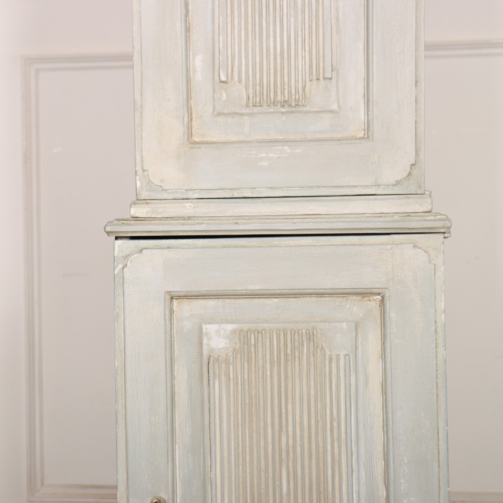 A tall, narrow Gustavian style two-tier painted wooden cabinet in pale grey with fluted panel doors and tapered feet. View VI