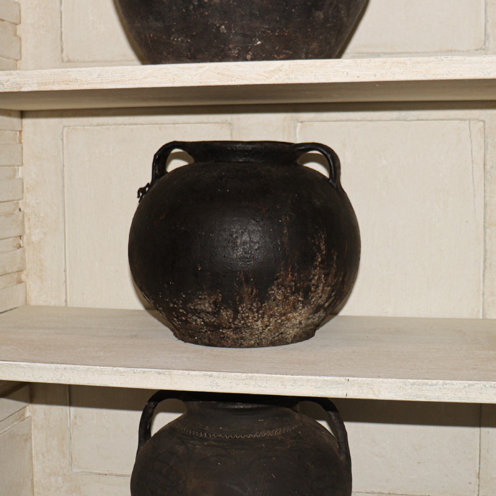 A collection of dark bulbous terracotta storage pots with small twin handles displayed on white shelving. View II