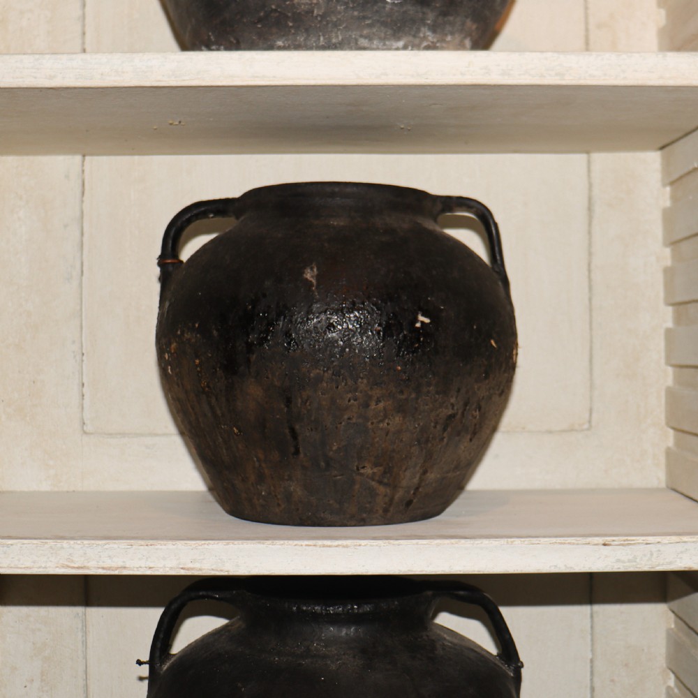 A collection of dark bulbous terracotta storage pots with small twin handles displayed on white shelving. View IV