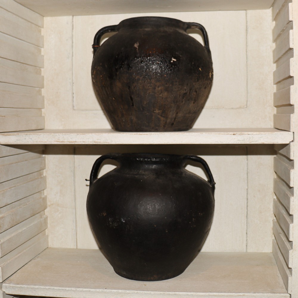 A collection of dark bulbous terracotta storage pots with small twin handles displayed on white shelving. View VIII