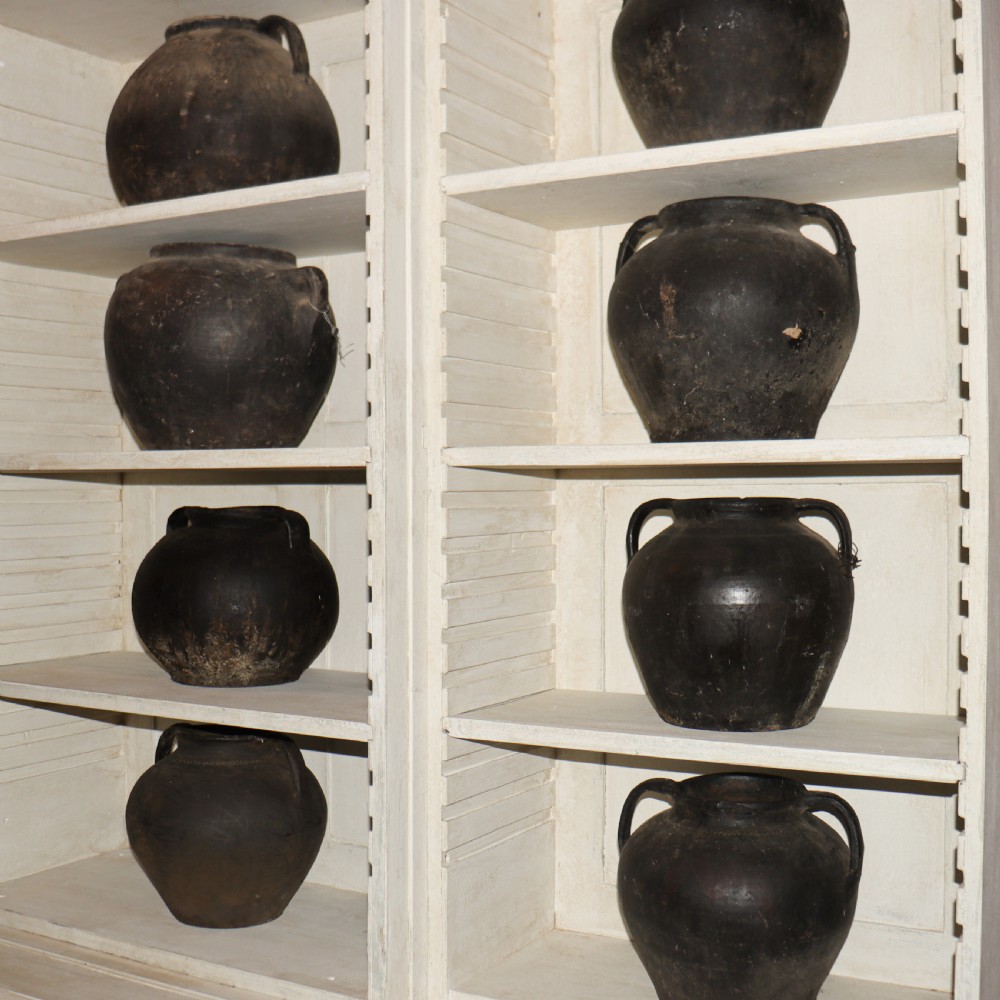 A collection of dark bulbous terracotta storage pots with small twin handles displayed on white shelving. View IX