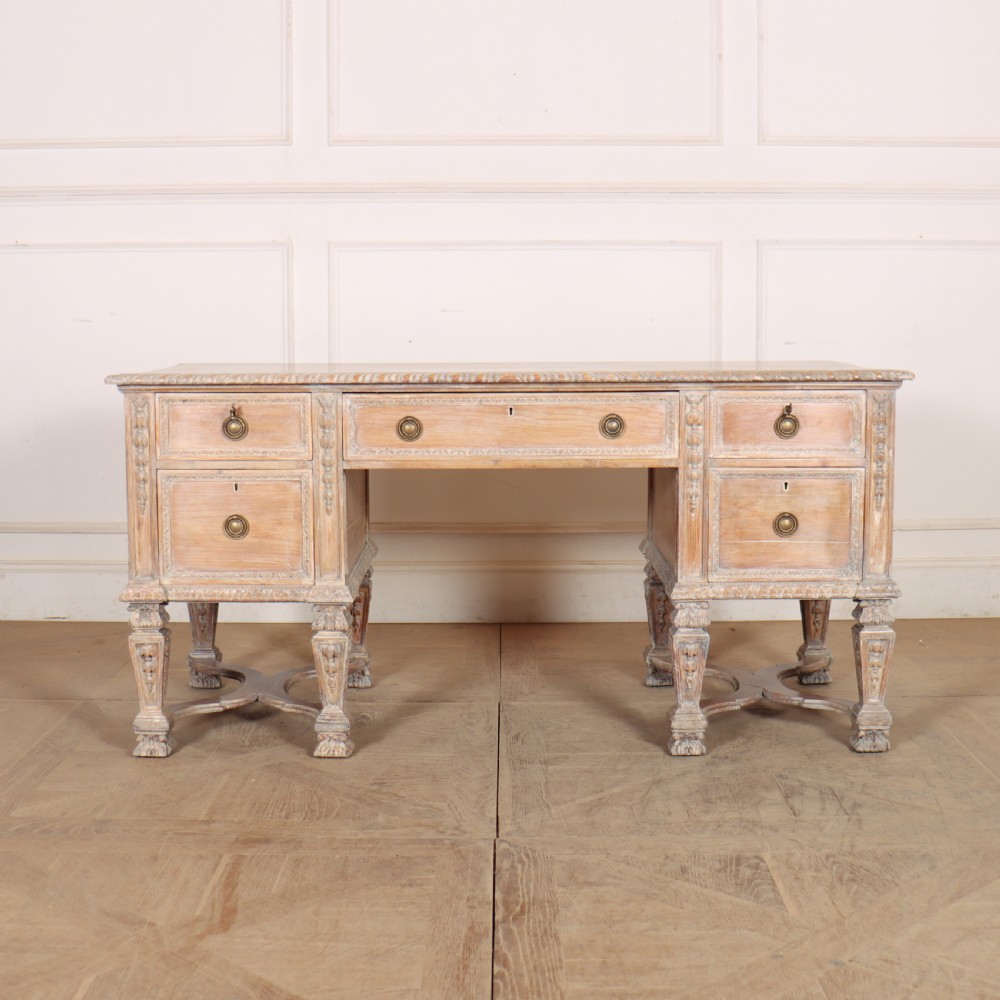A pale bleached oak kneehole writing desk with intricate carved legs, five drawers with brass drop handles, and double X-frame stretchers.