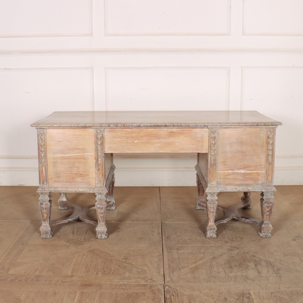 A pale bleached oak kneehole writing desk with intricate carved legs, five drawers with brass drop handles, and double X-frame stretchers. View XIV