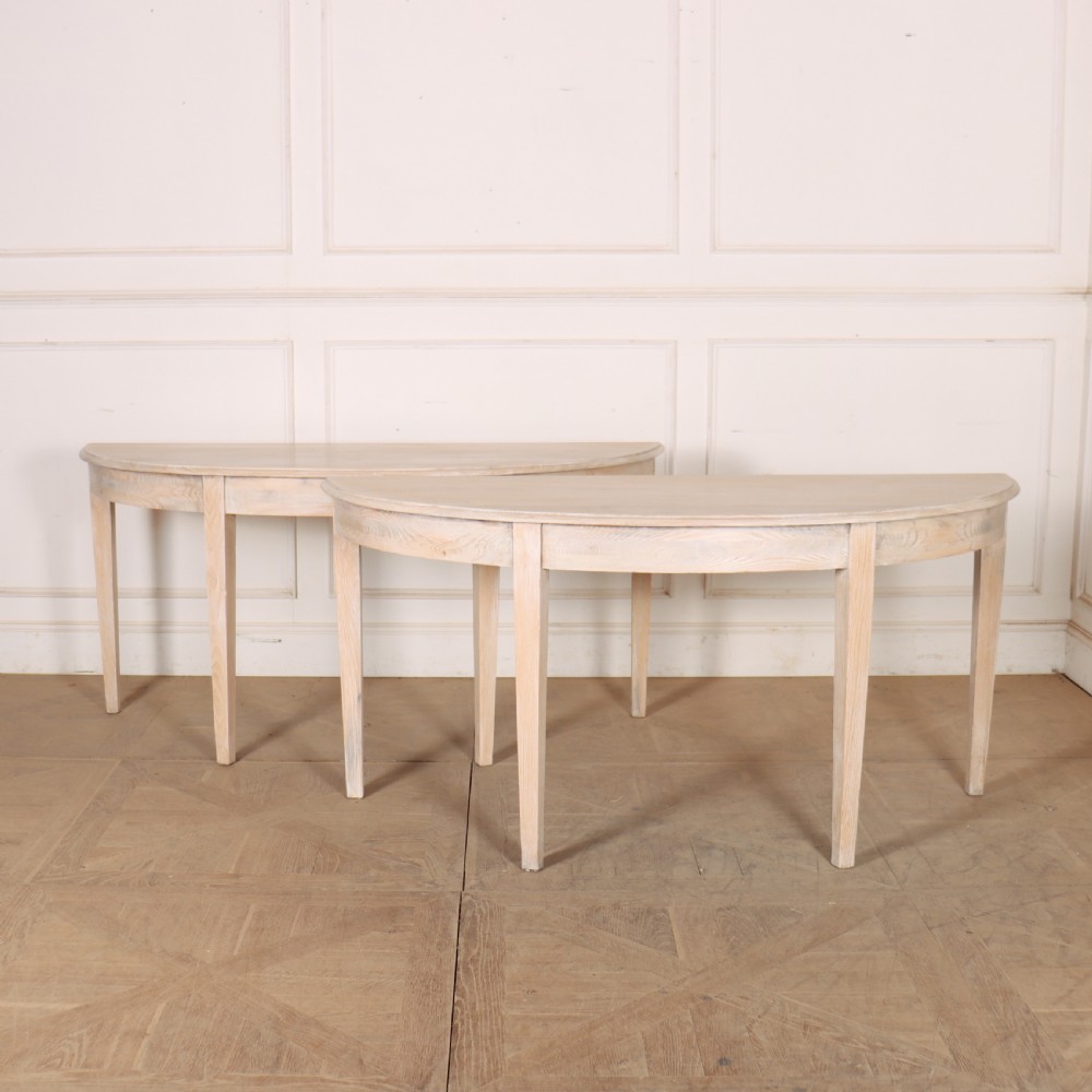 A pair of semi-circular, pale off-white painted console tables with tapered legs and flat backs designed to sit flush against a wall.