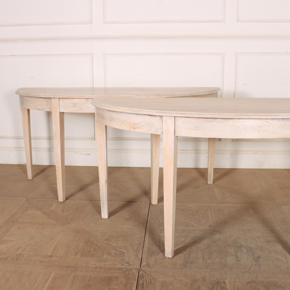 A pair of semi-circular, pale off-white painted console tables with tapered legs and flat backs designed to sit flush against a wall. View II