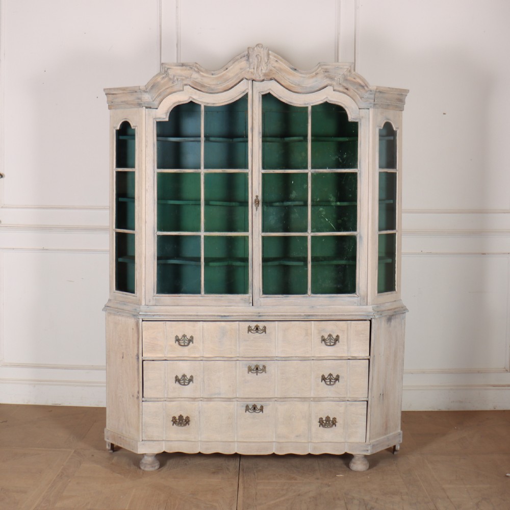An antique Dutch glazed display cabinet with a grey-washed exterior, green painted interior, curved cresting rail, and three drawers in the base.