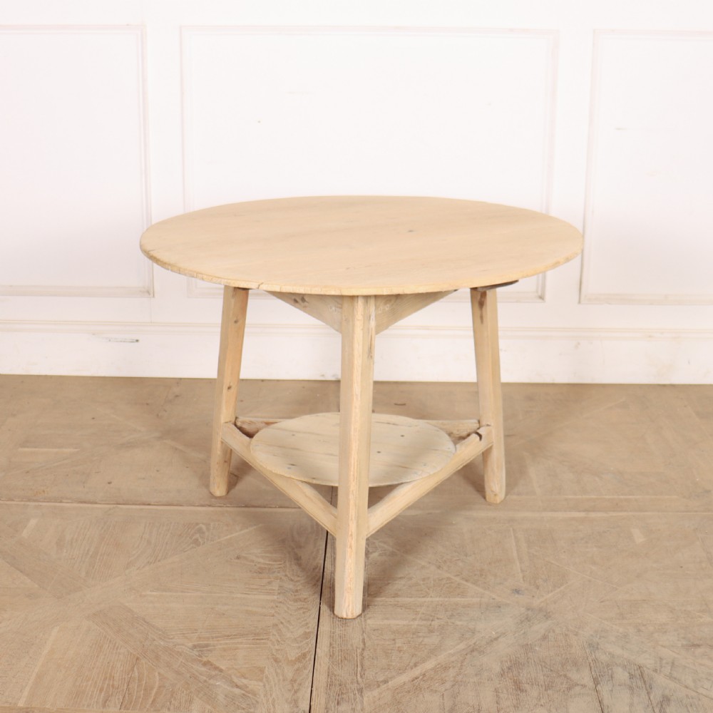 A round, pale wood three-legged cricket table with a lower triangular shelf resting on a parquet floor.