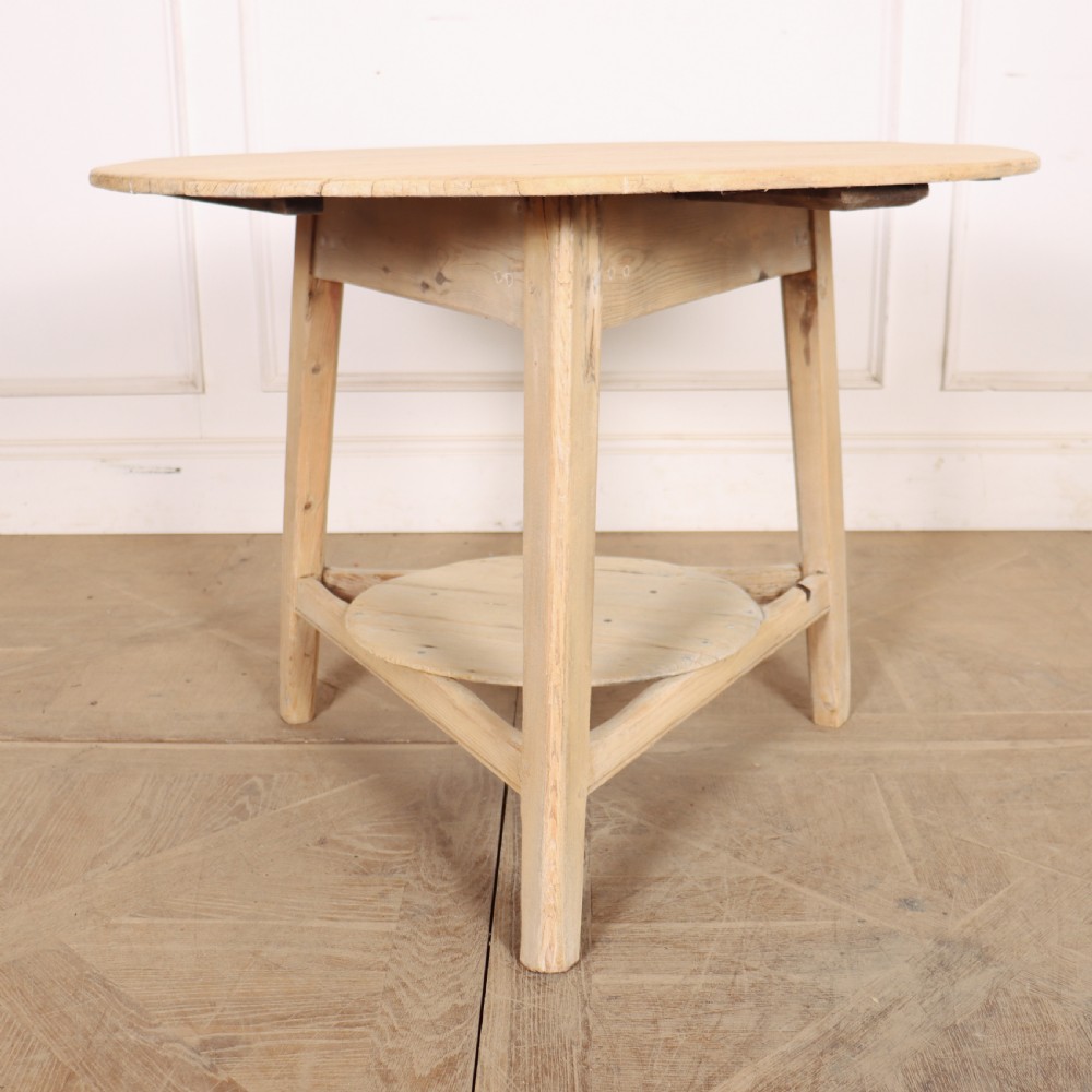 A round, pale wood three-legged cricket table with a lower triangular shelf resting on a parquet floor. View II