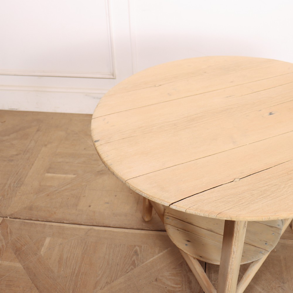 A round, pale wood three-legged cricket table with a lower triangular shelf resting on a parquet floor. View III