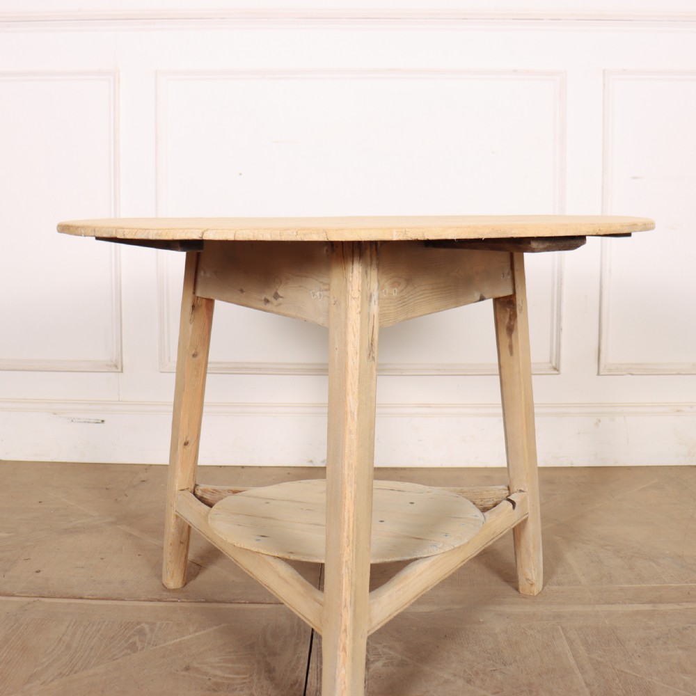 A round, pale wood three-legged cricket table with a lower triangular shelf resting on a parquet floor. View IV