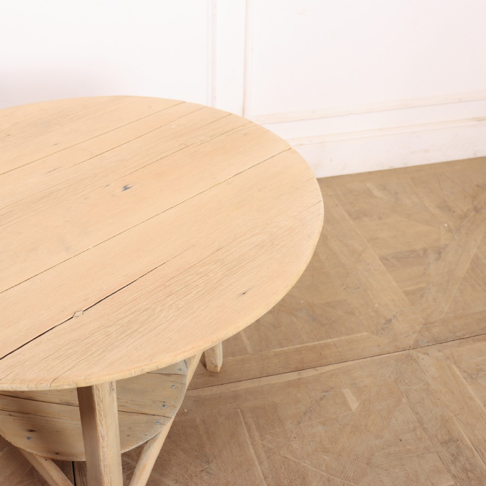 A round, pale wood three-legged cricket table with a lower triangular shelf resting on a parquet floor. View VI
