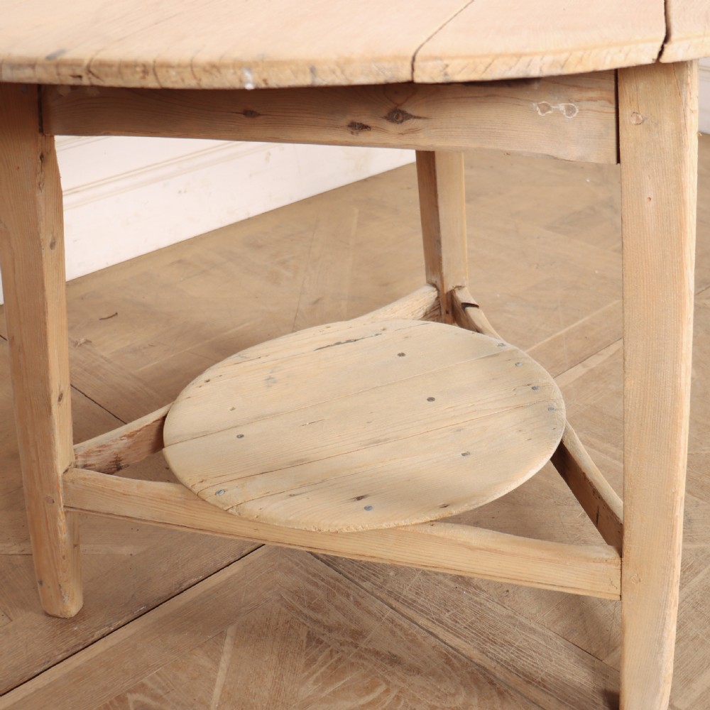 A round, pale wood three-legged cricket table with a lower triangular shelf resting on a parquet floor. View VII