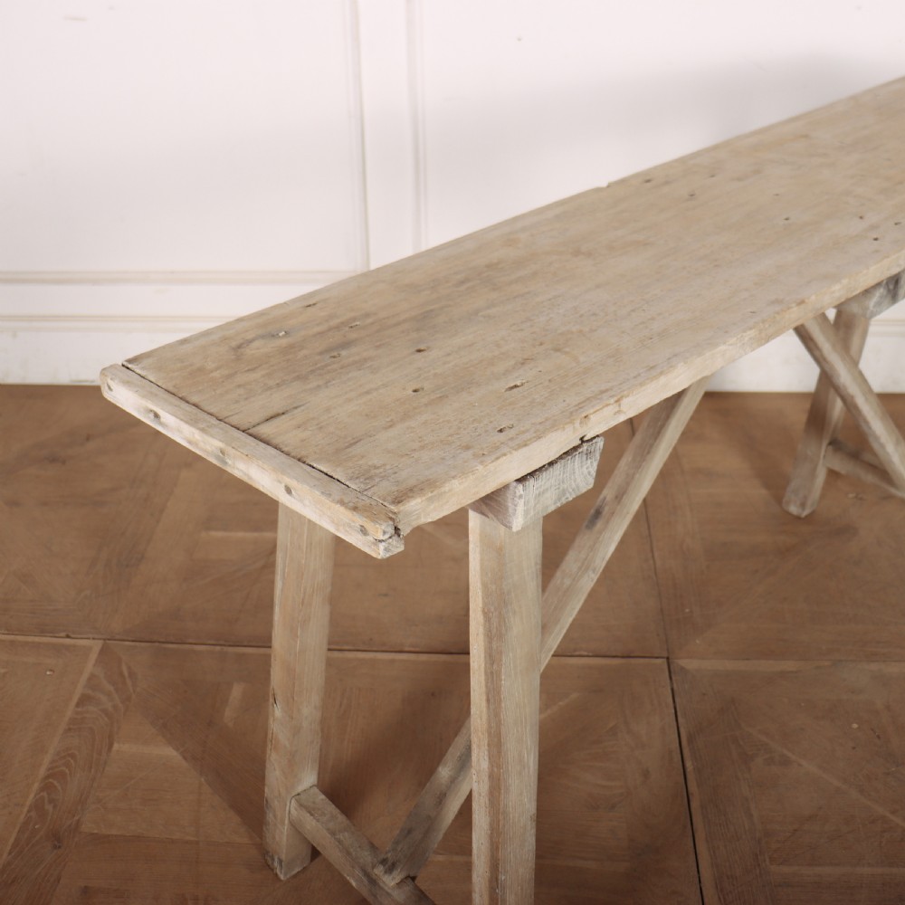 A long, narrow antique French rustic wooden trestle table with a weathered pale finish and diagonal cross-bracing. View II