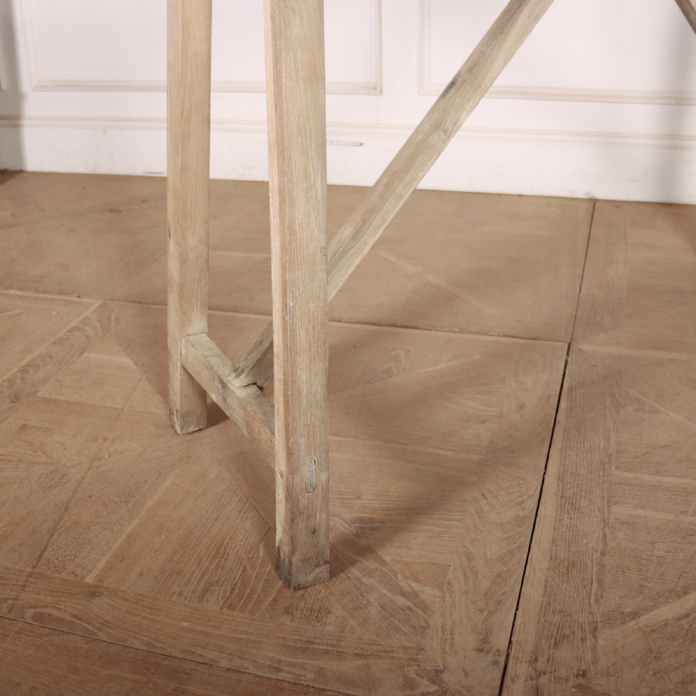 A long, narrow antique French rustic wooden trestle table with a weathered pale finish and diagonal cross-bracing. View III