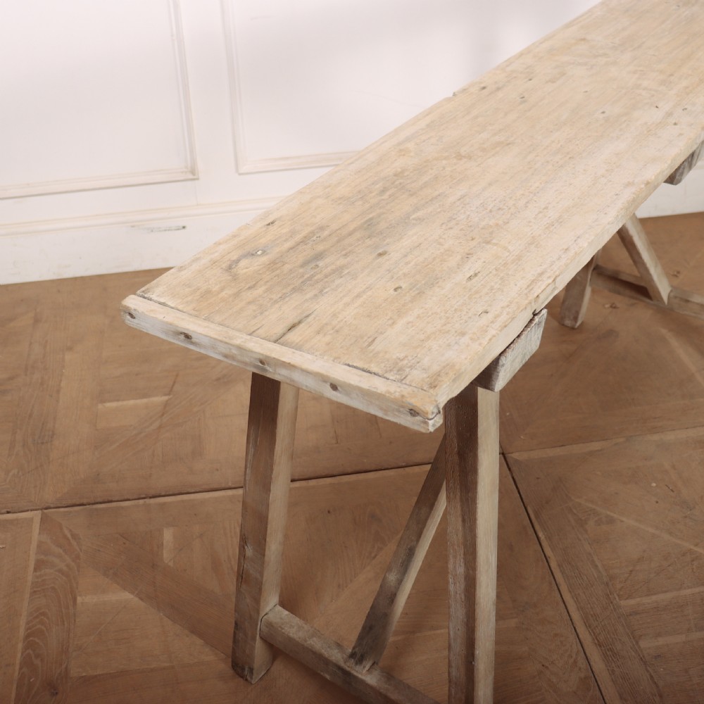 A long, narrow antique French rustic wooden trestle table with a weathered pale finish and diagonal cross-bracing. View VI