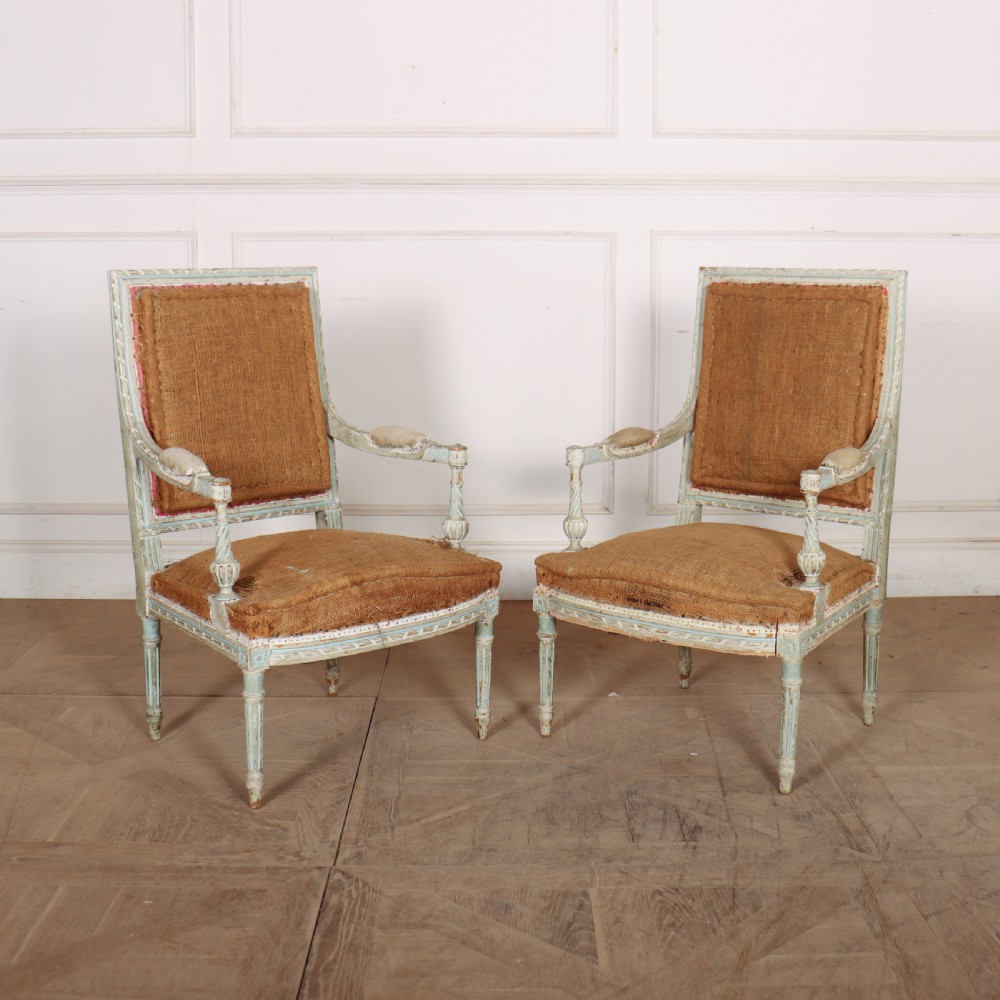 A pair of antique French Louis XVI open armchairs featuring carved, distressed light blue painted frames, fluted legs, and exposed hessian upholstery.