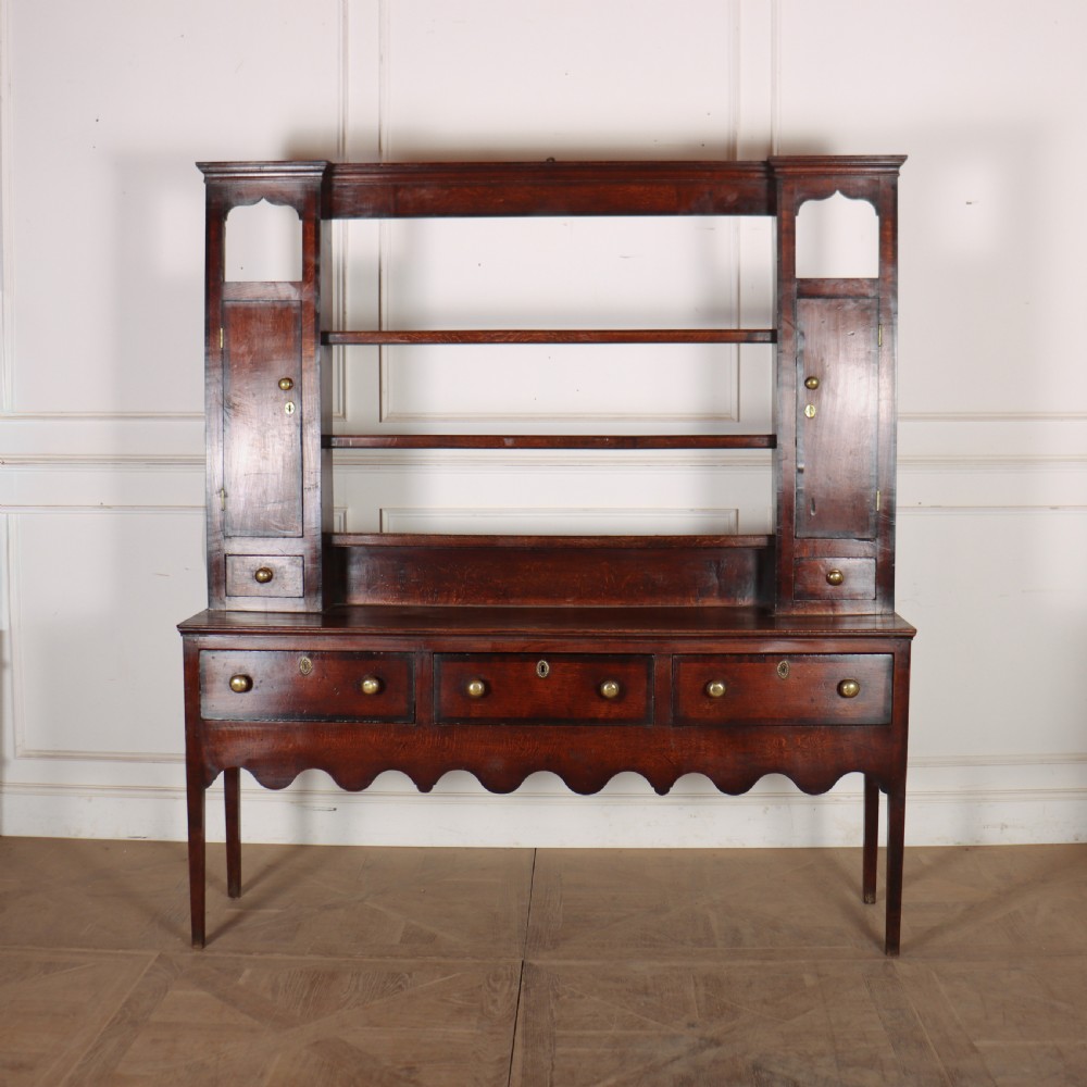 An 18th-century dark oak dresser featuring an open upper plate rack flanked by narrow side cupboards over a base with three drawers and a scalloped apron.