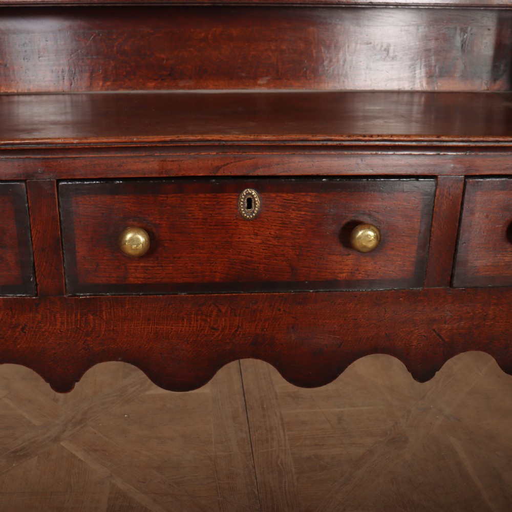An 18th-century dark oak dresser featuring an open upper plate rack flanked by narrow side cupboards over a base with three drawers and a scalloped apron. View VII
