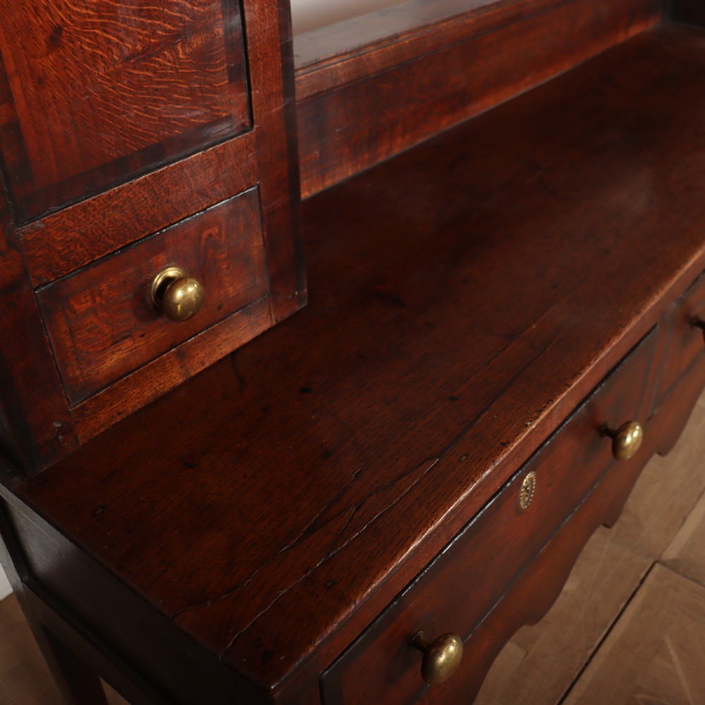 An 18th-century dark oak dresser featuring an open upper plate rack flanked by narrow side cupboards over a base with three drawers and a scalloped apron. View XII