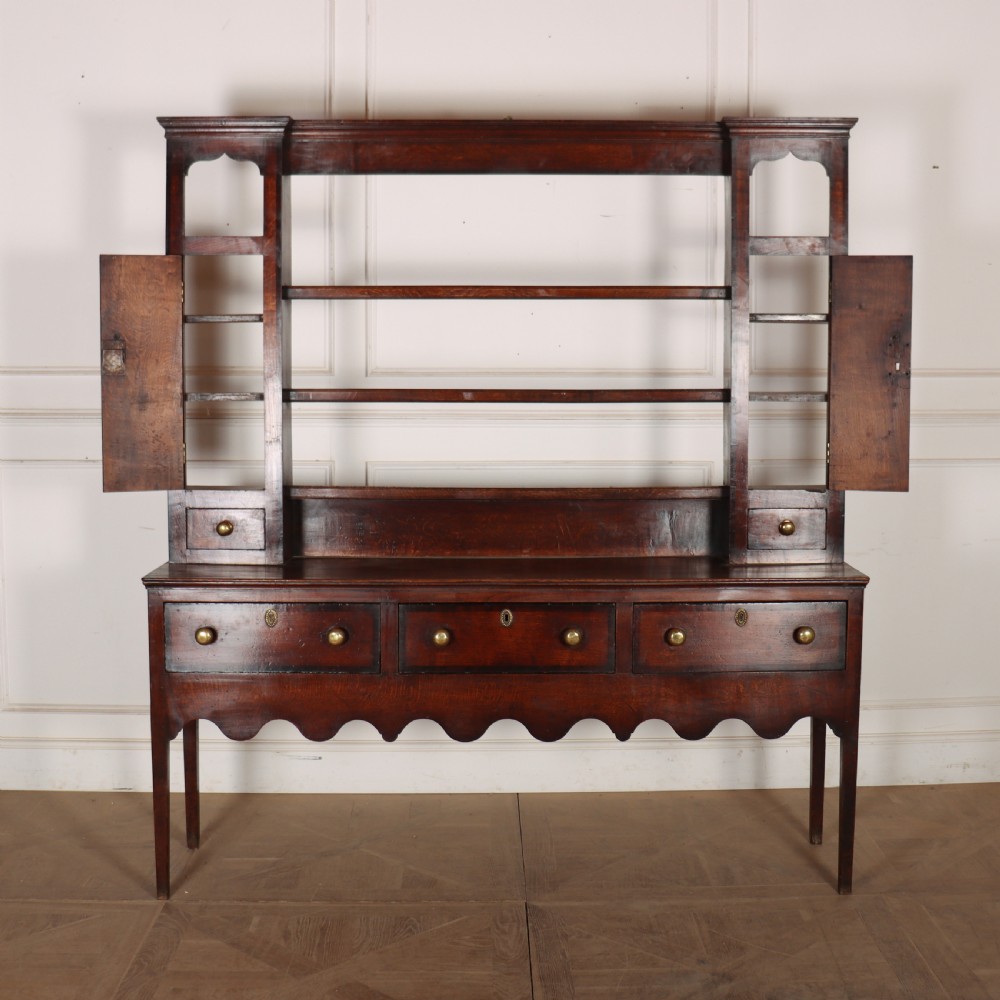 An 18th-century dark oak dresser featuring an open upper plate rack flanked by narrow side cupboards over a base with three drawers and a scalloped apron. View XIV