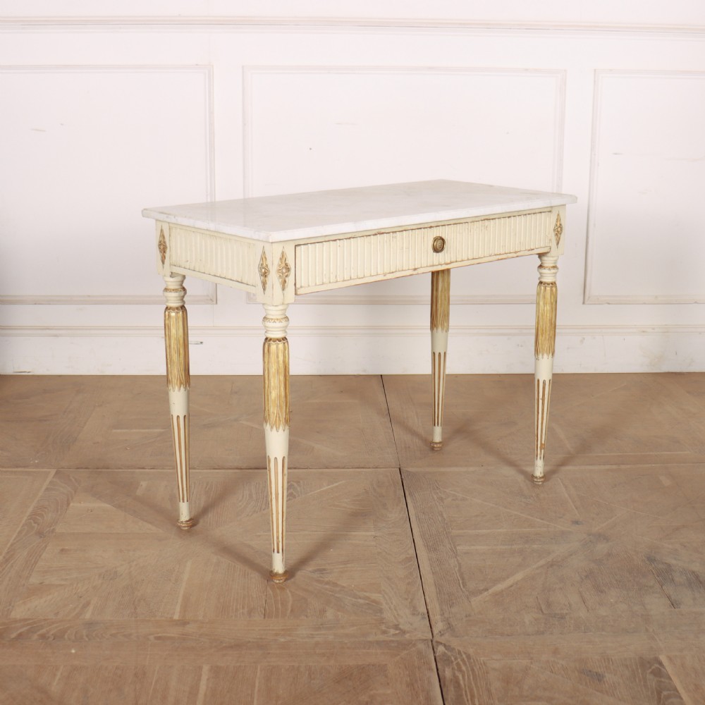 An off-white painted Swedish side table featuring a white marble top, fluted frieze with a central drawer knob, and tapered fluted legs with gilded accents.