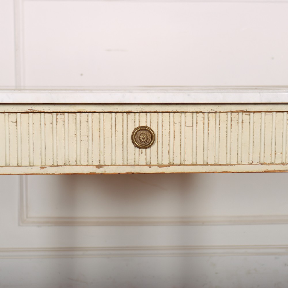 An off-white painted Swedish side table featuring a white marble top, fluted frieze with a central drawer knob, and tapered fluted legs with gilded accents. View VII