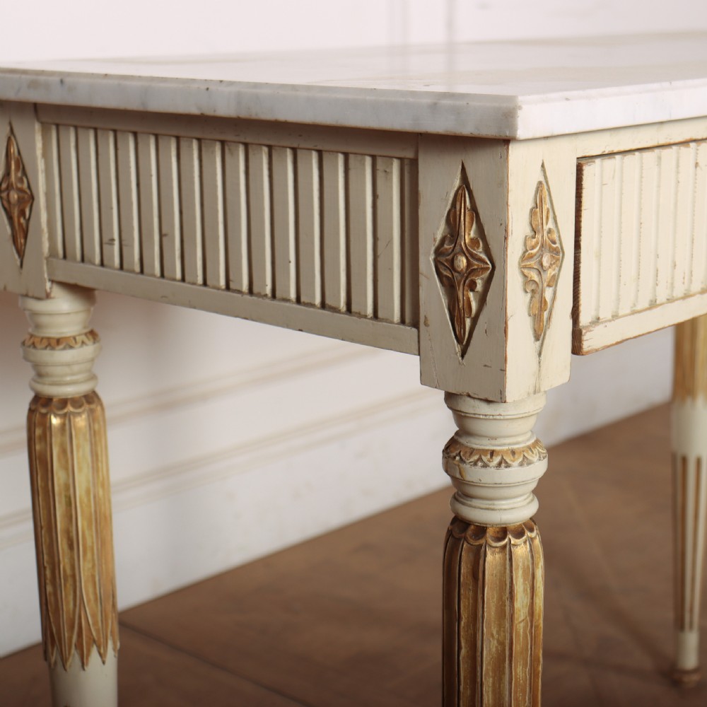 An off-white painted Swedish side table featuring a white marble top, fluted frieze with a central drawer knob, and tapered fluted legs with gilded accents. View X