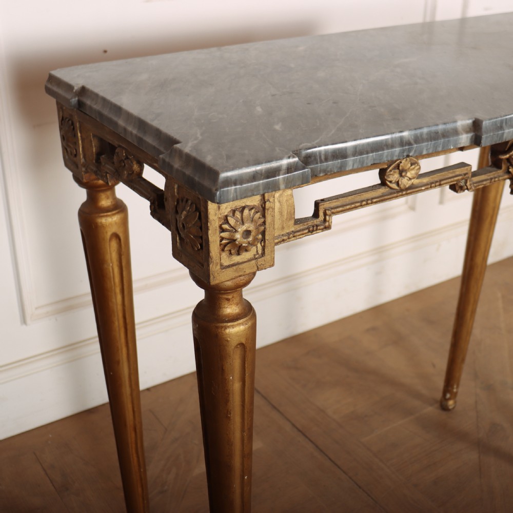 A rectangular gilded console table featuring a grey marble top, carved rams head central motif, rosettes, and tapered legs. View III
