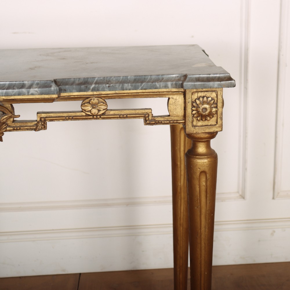 A rectangular gilded console table featuring a grey marble top, carved rams head central motif, rosettes, and tapered legs. View IV