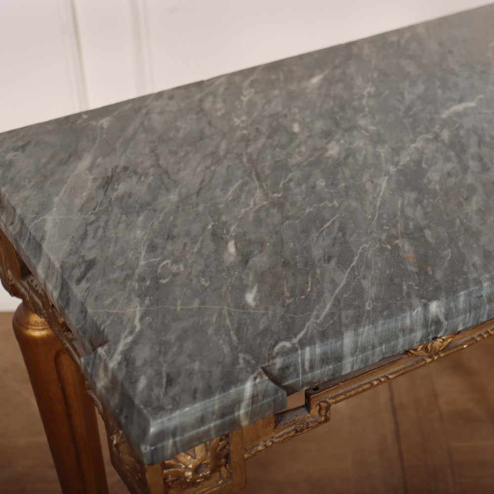 A rectangular gilded console table featuring a grey marble top, carved rams head central motif, rosettes, and tapered legs. View IX