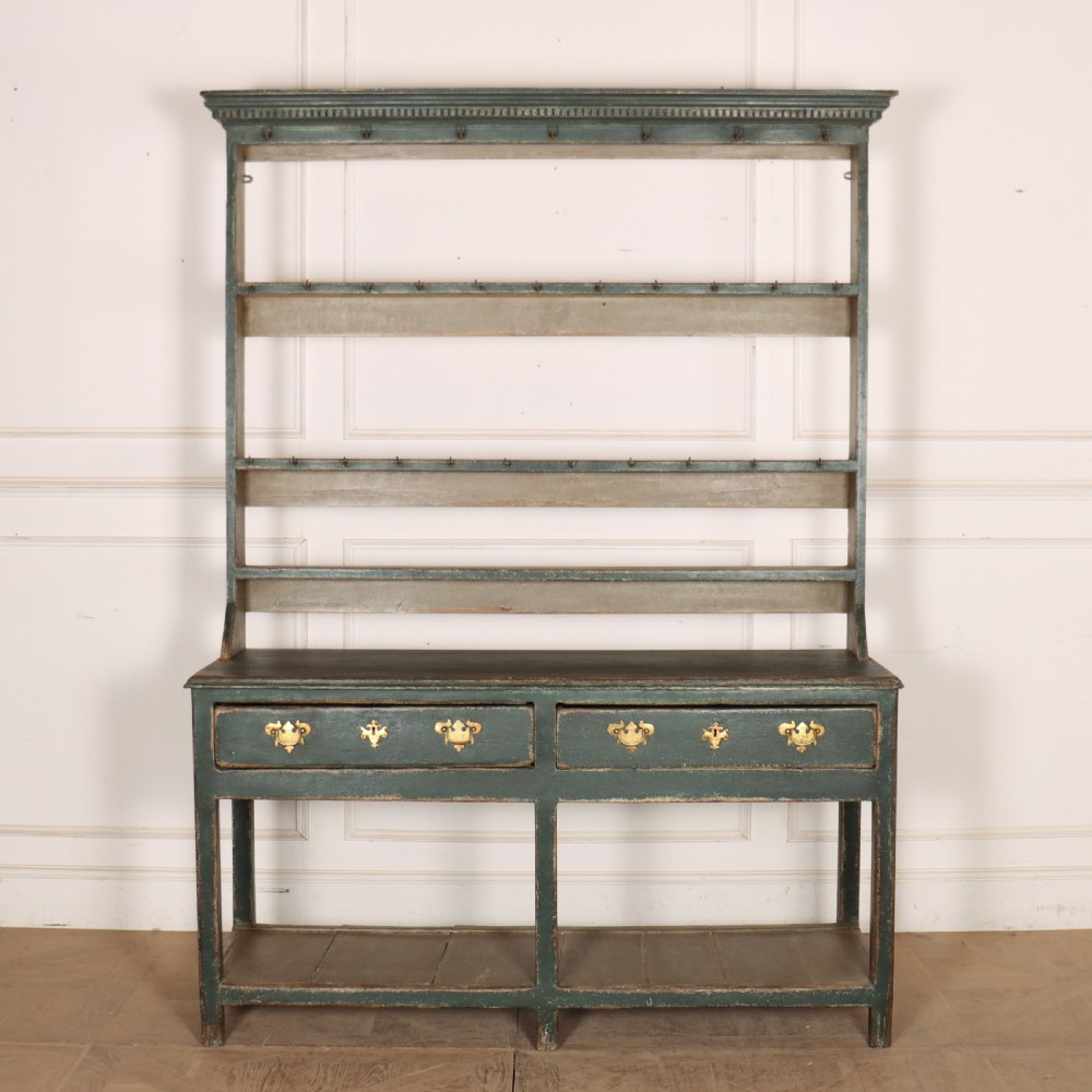 An antique dark green painted wooden pot board dresser featuring an upper three-tier plate rack with cup hooks above two drawers and an open bottom shelf.