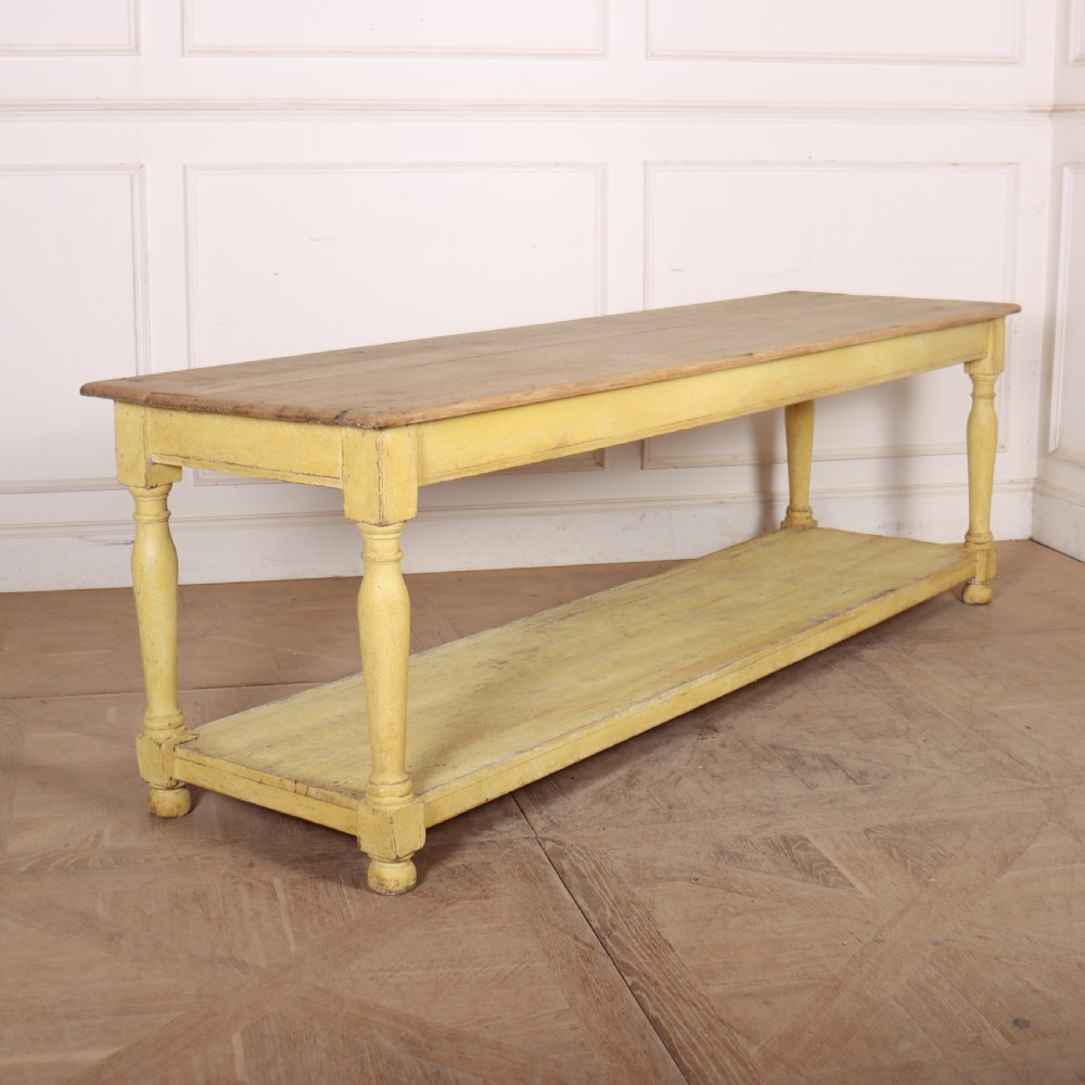 A long French drapers table featuring a natural wood plank top, a yellow painted apron and turned legs, and a low potboard shelf.