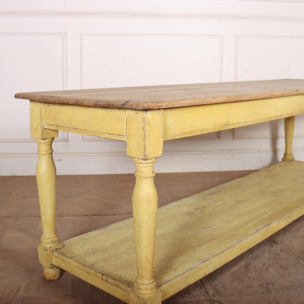 A long French drapers table featuring a natural wood plank top, a yellow painted apron and turned legs, and a low potboard shelf. View II