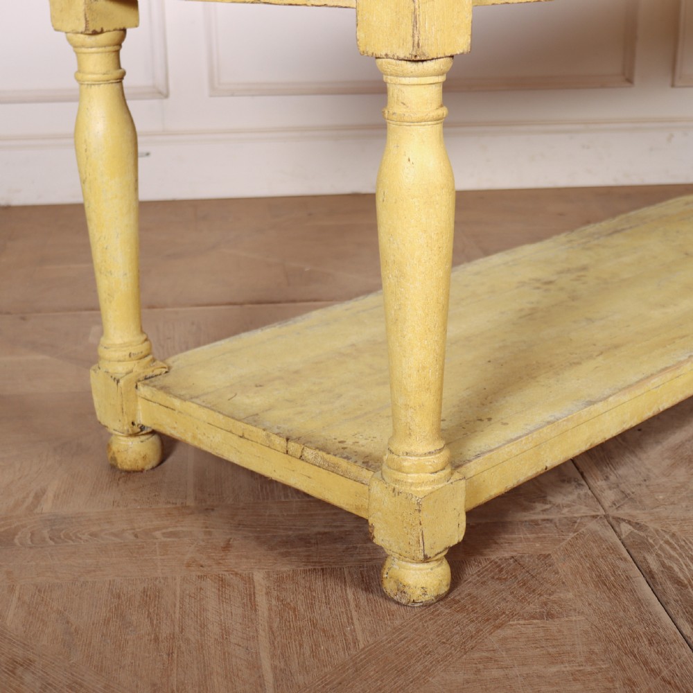 A long French drapers table featuring a natural wood plank top, a yellow painted apron and turned legs, and a low potboard shelf. View III
