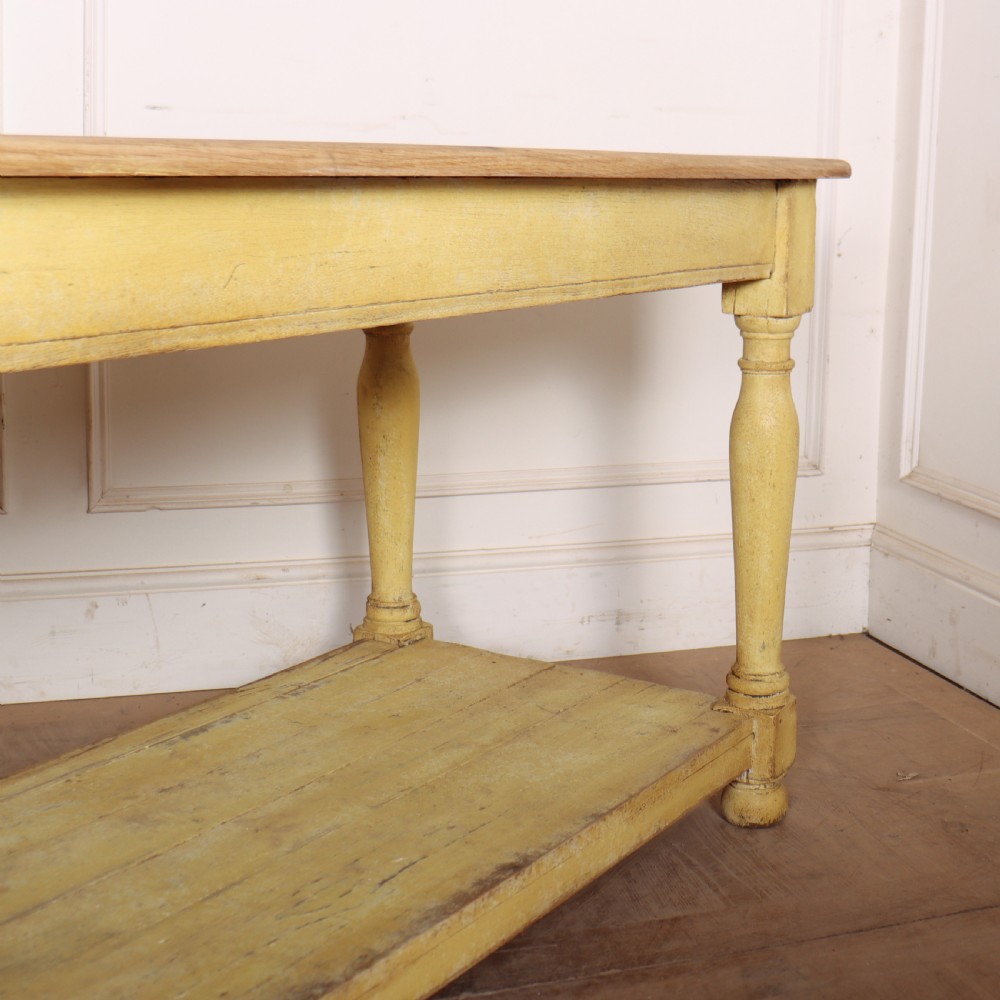 A long French drapers table featuring a natural wood plank top, a yellow painted apron and turned legs, and a low potboard shelf. View V