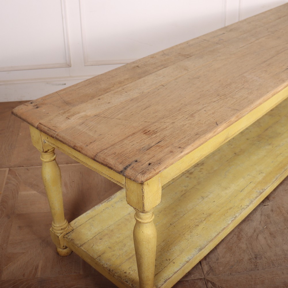 A long French drapers table featuring a natural wood plank top, a yellow painted apron and turned legs, and a low potboard shelf. View VII