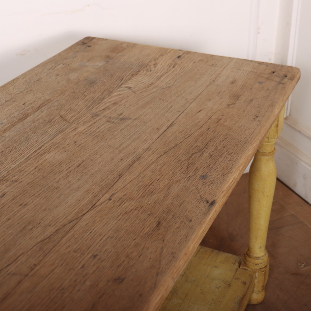 A long French drapers table featuring a natural wood plank top, a yellow painted apron and turned legs, and a low potboard shelf. View IX