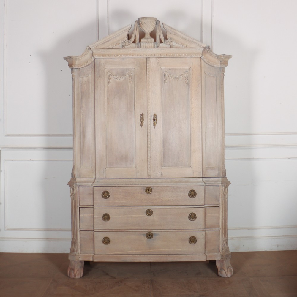 A Dutch bleached oak linen cupboard featuring a top pediment with a carved urn, two panelled doors decorated with carved swags, and three lower drawers resting on claw feet.