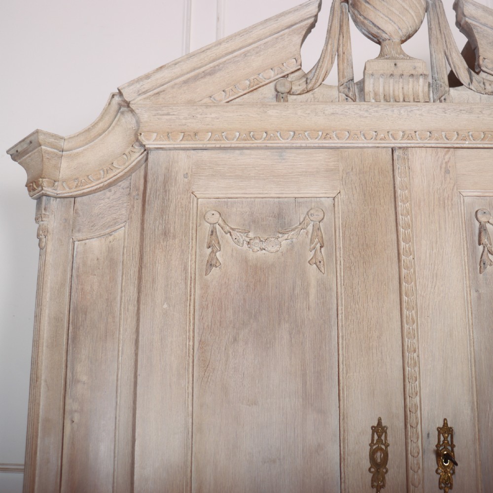 A Dutch bleached oak linen cupboard featuring a top pediment with a carved urn, two panelled doors decorated with carved swags, and three lower drawers resting on claw feet. View IV