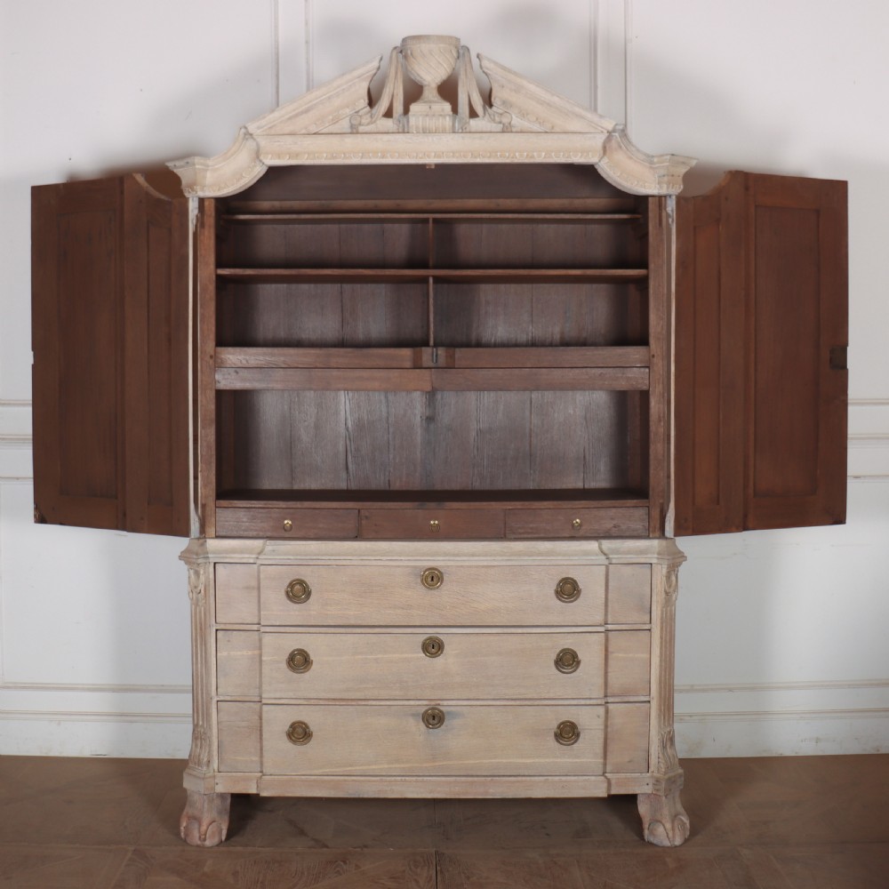 A Dutch bleached oak linen cupboard featuring a top pediment with a carved urn, two panelled doors decorated with carved swags, and three lower drawers resting on claw feet. View VII