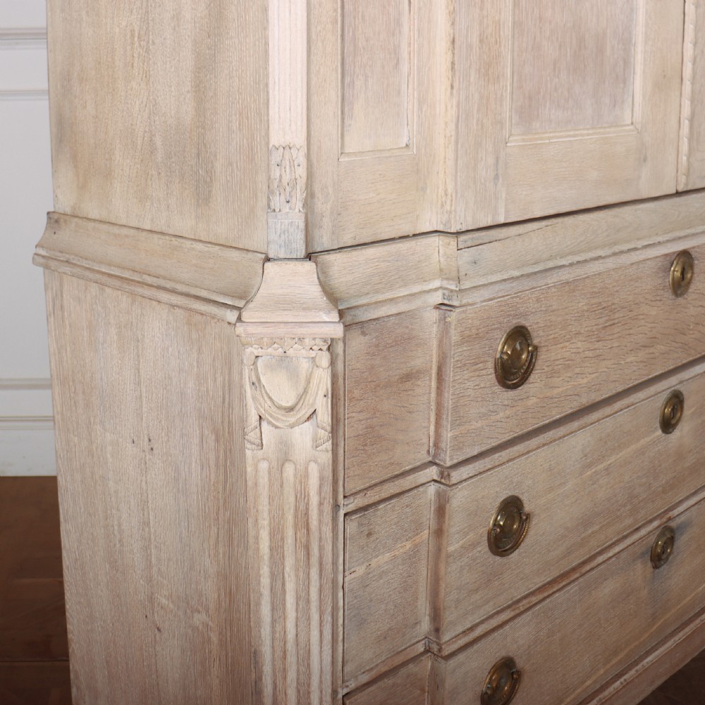 A Dutch bleached oak linen cupboard featuring a top pediment with a carved urn, two panelled doors decorated with carved swags, and three lower drawers resting on claw feet. View X