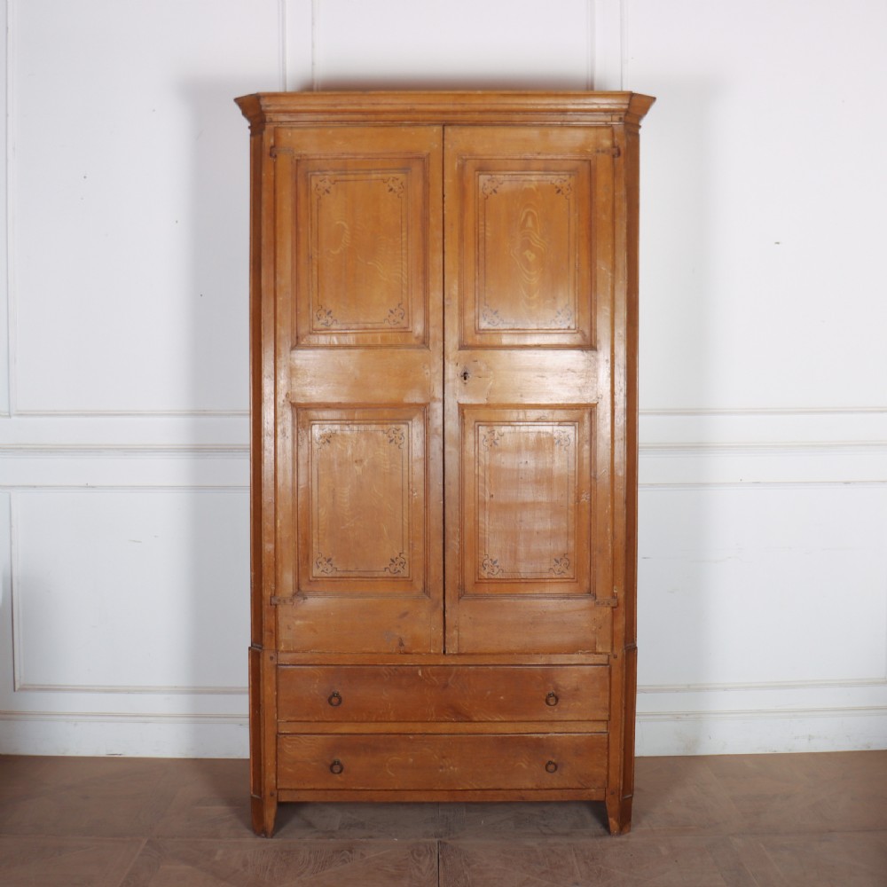An antique freestanding pine linen cupboard featuring two panelled upper doors decorated with painted corner flourishes above two full-width lower drawers.