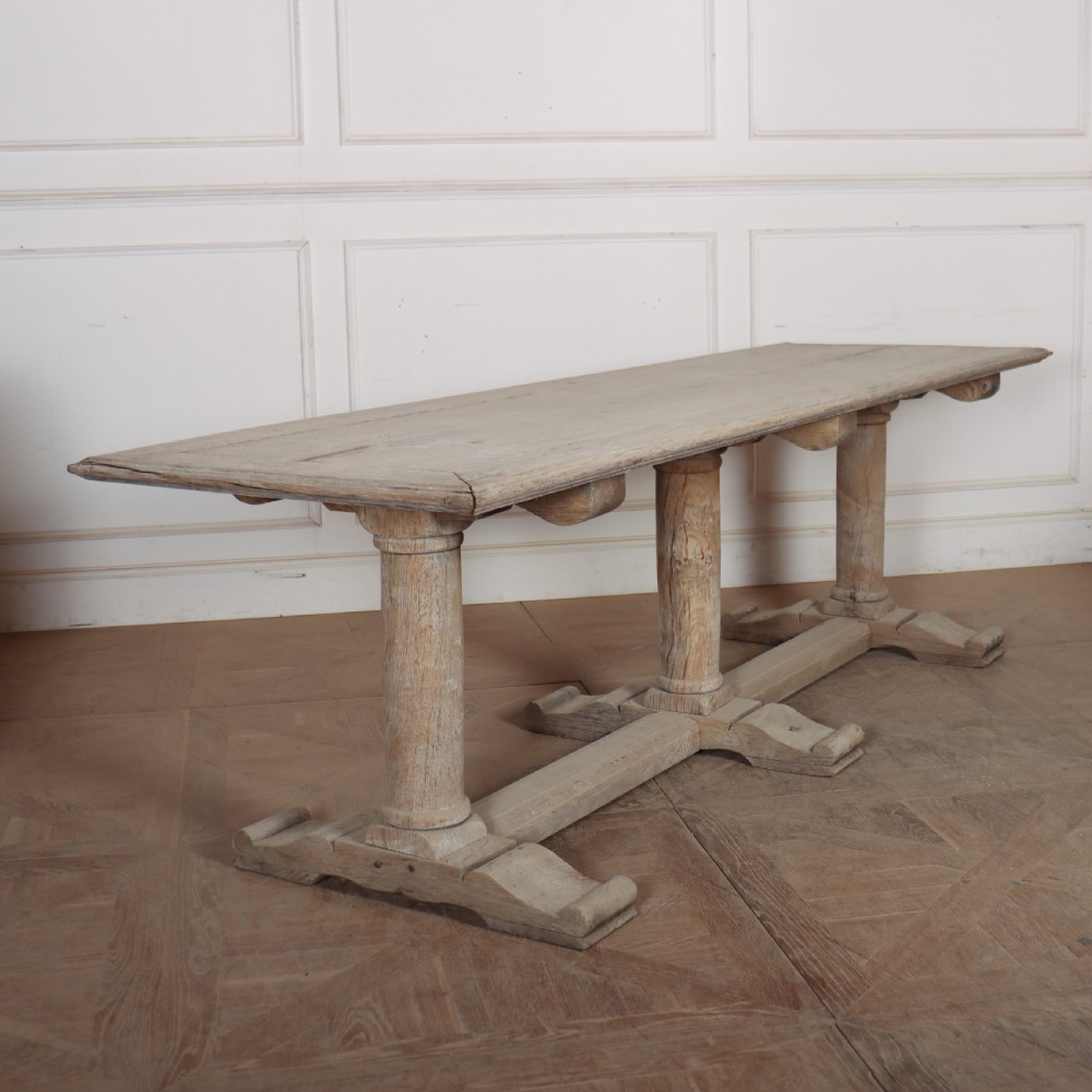 A long rectangular bleached oak trestle table featuring three column support legs and scrolled feet.
