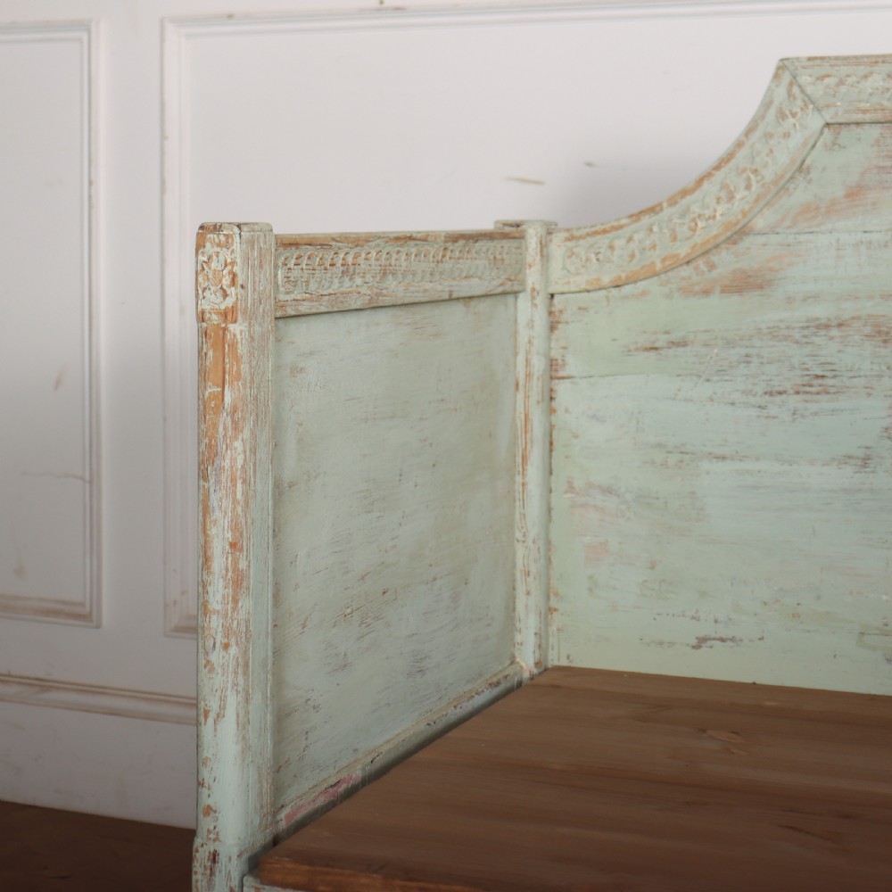 A pale green painted Swedish wooden bench featuring a shaped crest rail, panelled sides, and a natural pine seat. View II