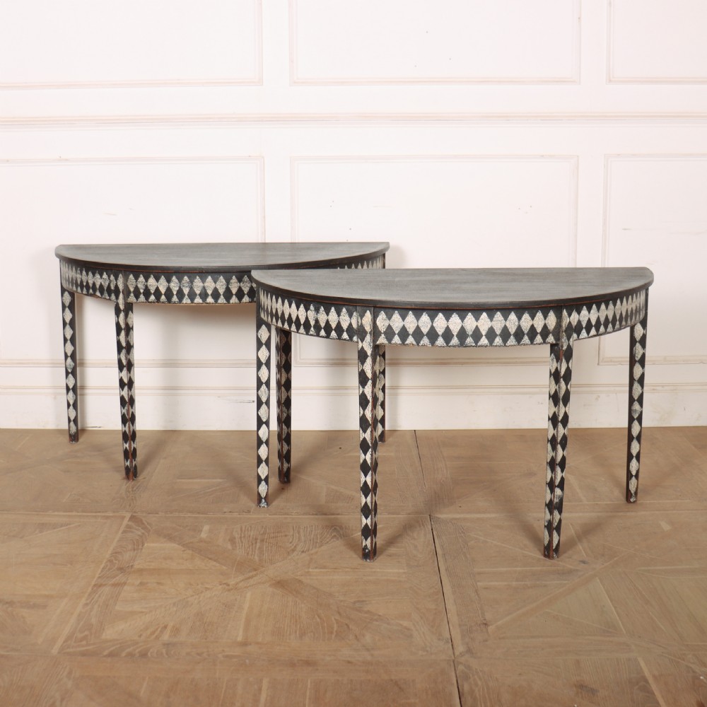 A pair of semi-circular painted timber console tables decorated with a black and white diamond harlequin pattern along the aprons and tapered legs.