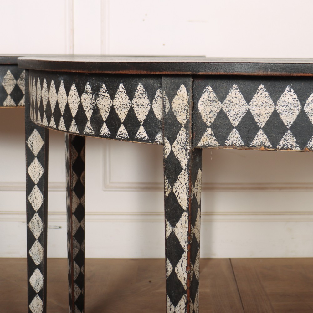 A pair of semi-circular painted timber console tables decorated with a black and white diamond harlequin pattern along the aprons and tapered legs. View V