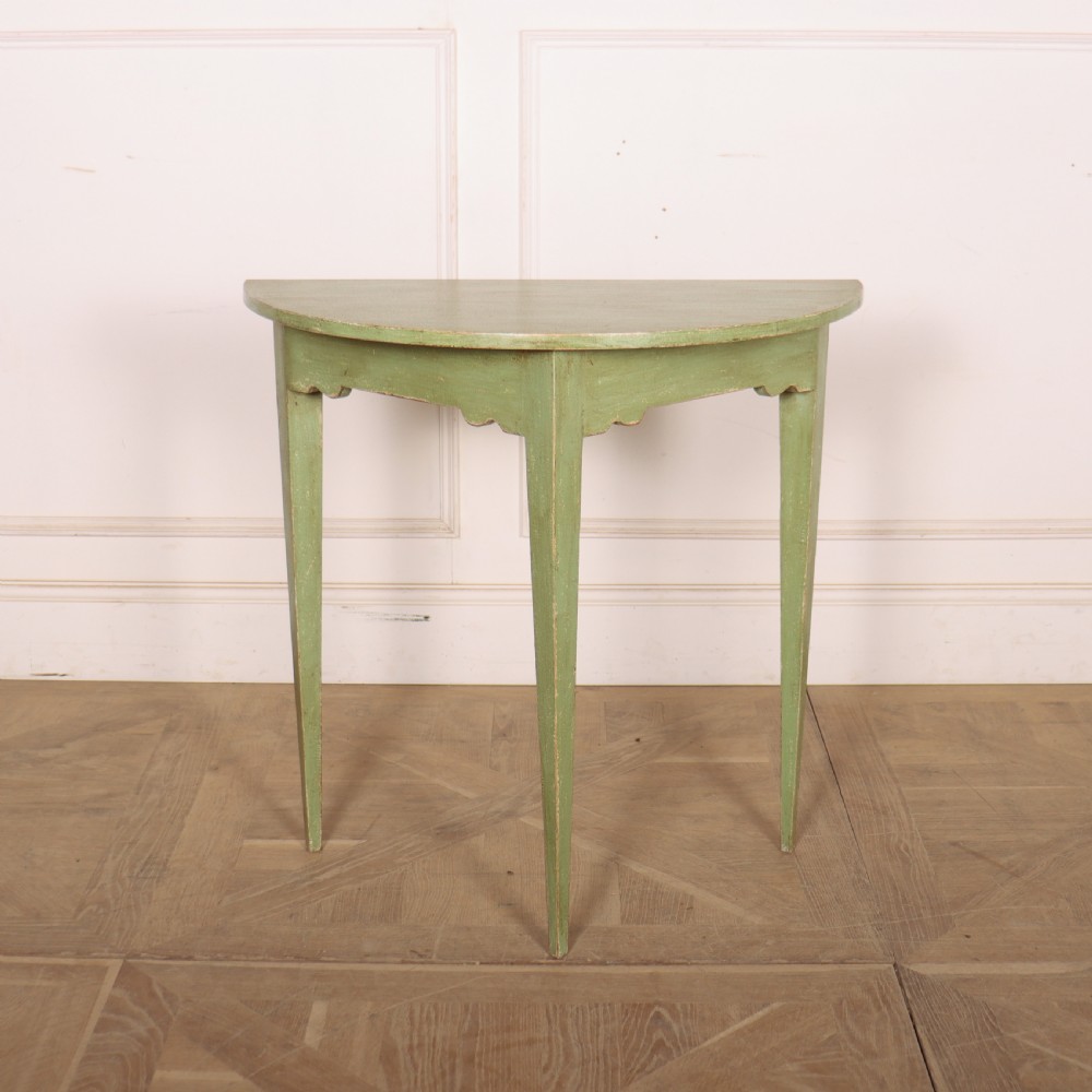 A semi-circular green painted wooden console table featuring three tapered legs and a scalloped apron against a white wall.